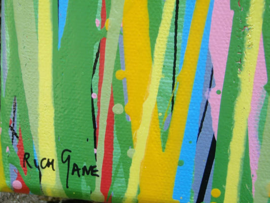 A colourful abstract painting of a grassy field with splattered flowers in orange, yellow, blue, and white against a light blue sky, hand-painted on to a large framed 100 by 70 cm canvas - detail