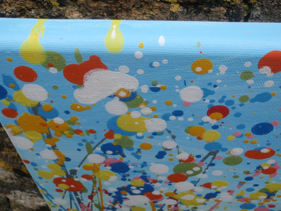 A colourful abstract painting of a grassy field with tall green, yellow, and red strokes, dotted with red, blue, orange, and pink flowers, beneath a light blue sky with white paint splatter. Hand-painted on to a large 100 by 70 cm canvas - detail
