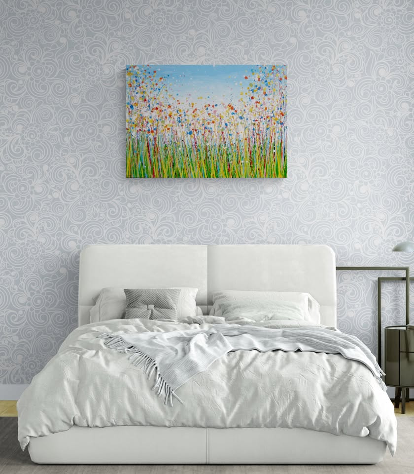 A colourful abstract painting of a grassy field with tall green, yellow, and red strokes, dotted with red, blue, orange, and pink flowers, beneath a light blue sky with white paint splatter. Hand-painted on to a large 100 by 70 cm canvas - shown on a wall in a bedroom