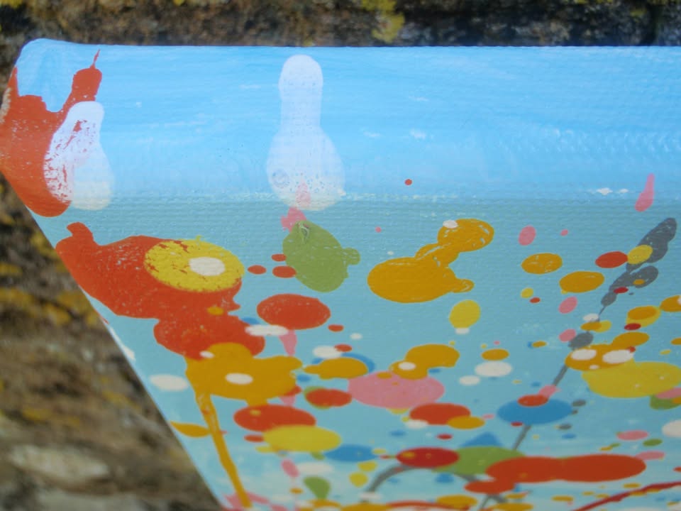 A colourful abstract painting of a grassy field with splattered flowers in orange, yellow, blue, and white against a light blue sky, hand-painted on to a large framed 100 by 70 cm canvas - detail