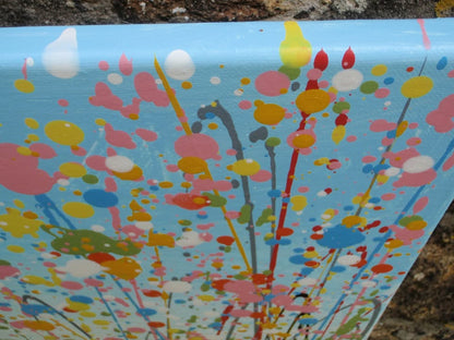 A colourful abstract painting of a grassy field under a blue sky, with splashes of pink, yellow, blue, green, and orange dots and lines throughout, hand-painted on to a large canvas - detail