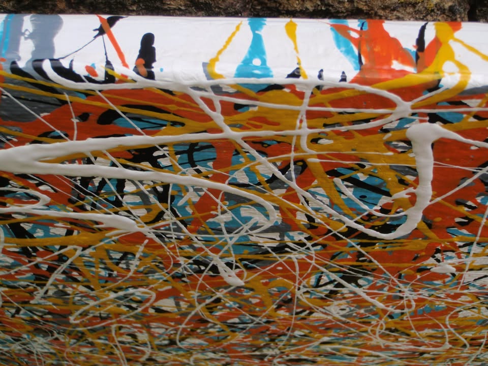 An abstract painting filled with dense, energetic splatters and drips in orange, yellow, white, black, and light blue, hand-painted on to a large framed wooden panel - detail