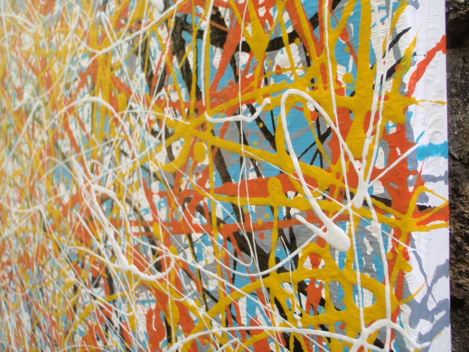 An abstract painting filled with dense, energetic splatters and drips in orange, yellow, white, black, and light blue, hand-painted on to a large framed wooden panel - detail